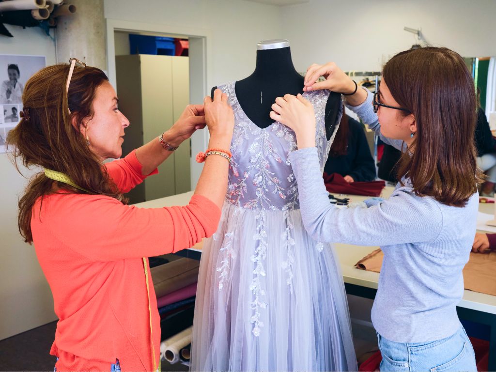 Die Werkstattleiterin mit Gesellin arbeiten an einem weißen, spitzenbesetzten Kleid